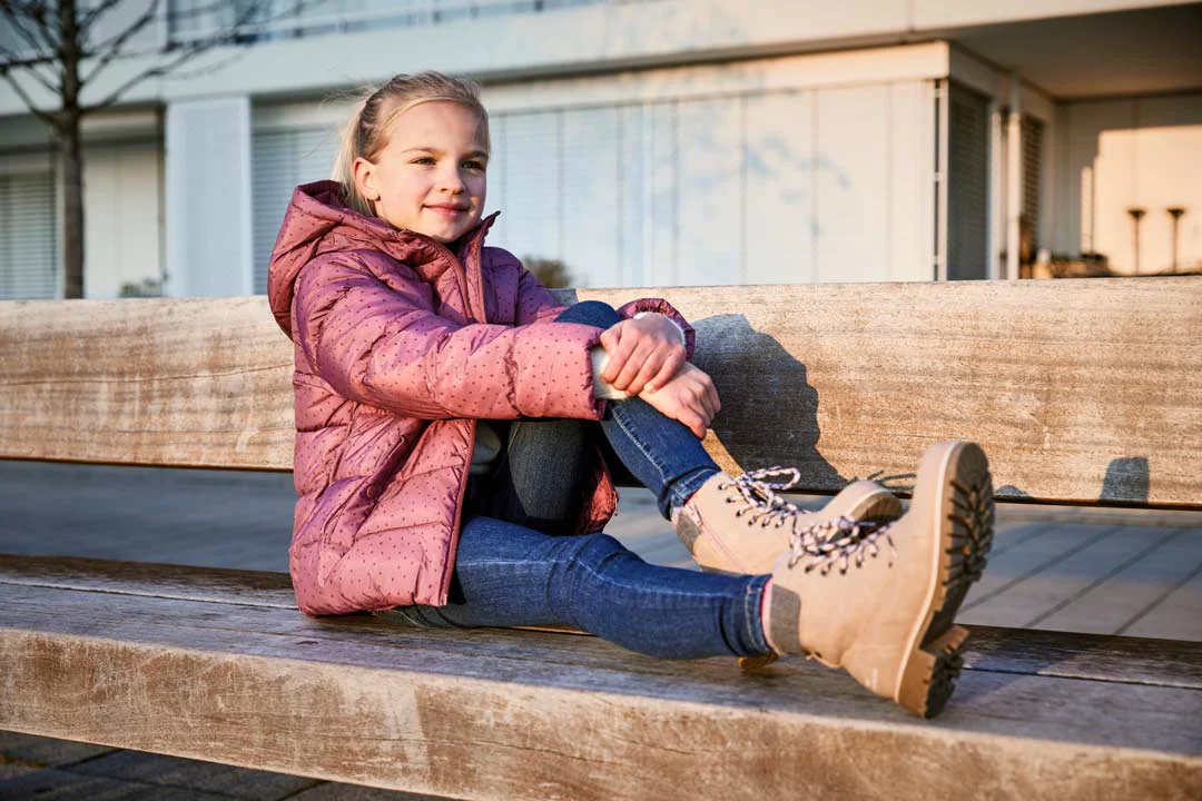 Kind sitzt auf einer Holzbank, trägt rosa Steppjacke, blaue Jeans und beige Schnürstiefel mit Profilsohle.