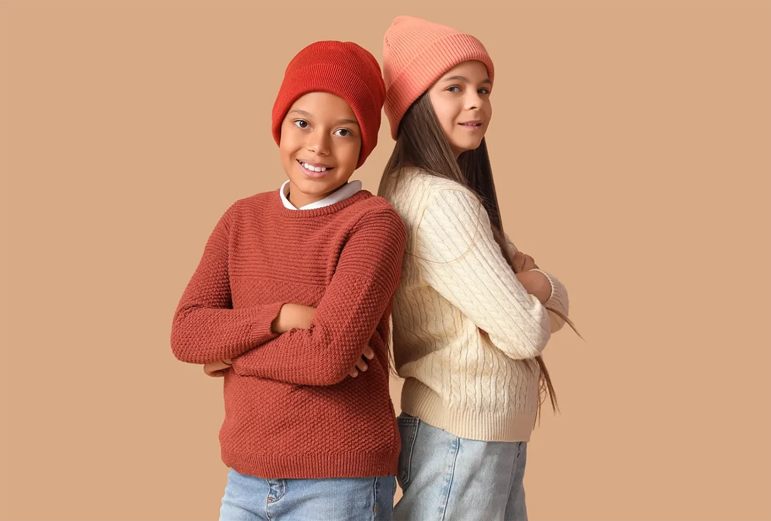 Zwei Kinder stehen Rücken an Rücken, beide mit verschränkten Armen, eines trägt roten Pullover und Mütze, das andere cremefarbenen Pullover und rosa Mütze.