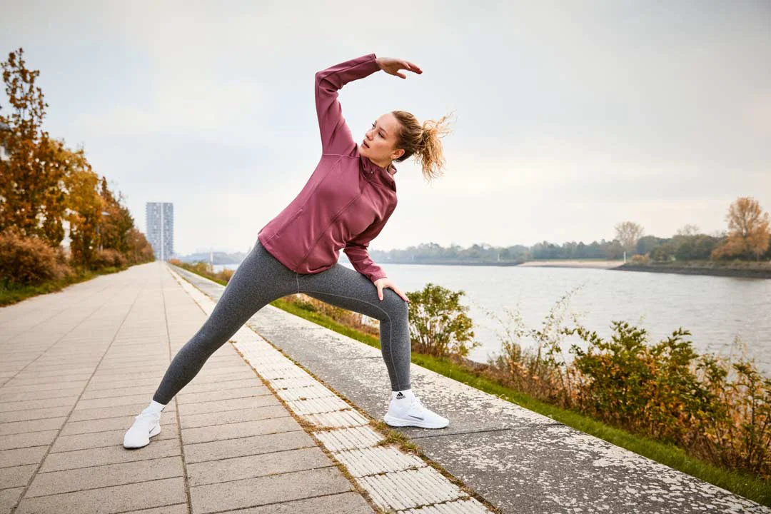 Frau in Sportkleidung dehnt sich seitlich auf einem Weg neben einem Fluss mit Bäumen im Hintergrund.