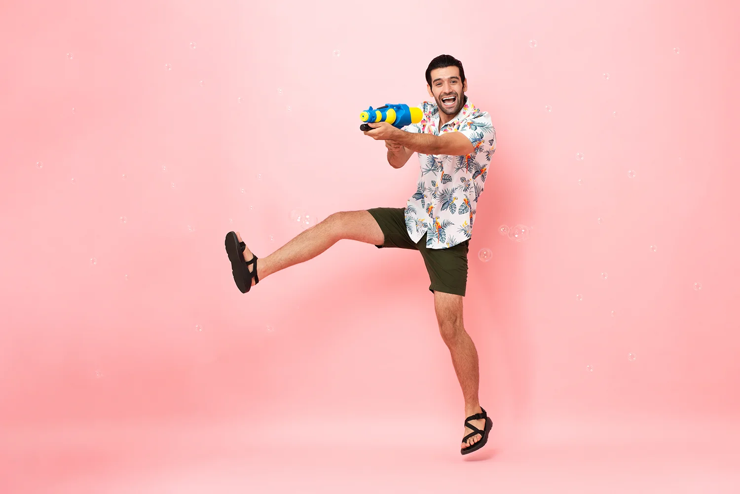 Mann in floralem Hemd, grünen Shorts und schwarzen Sandalen springt mit Wasserpistole vor rosa Hintergrund