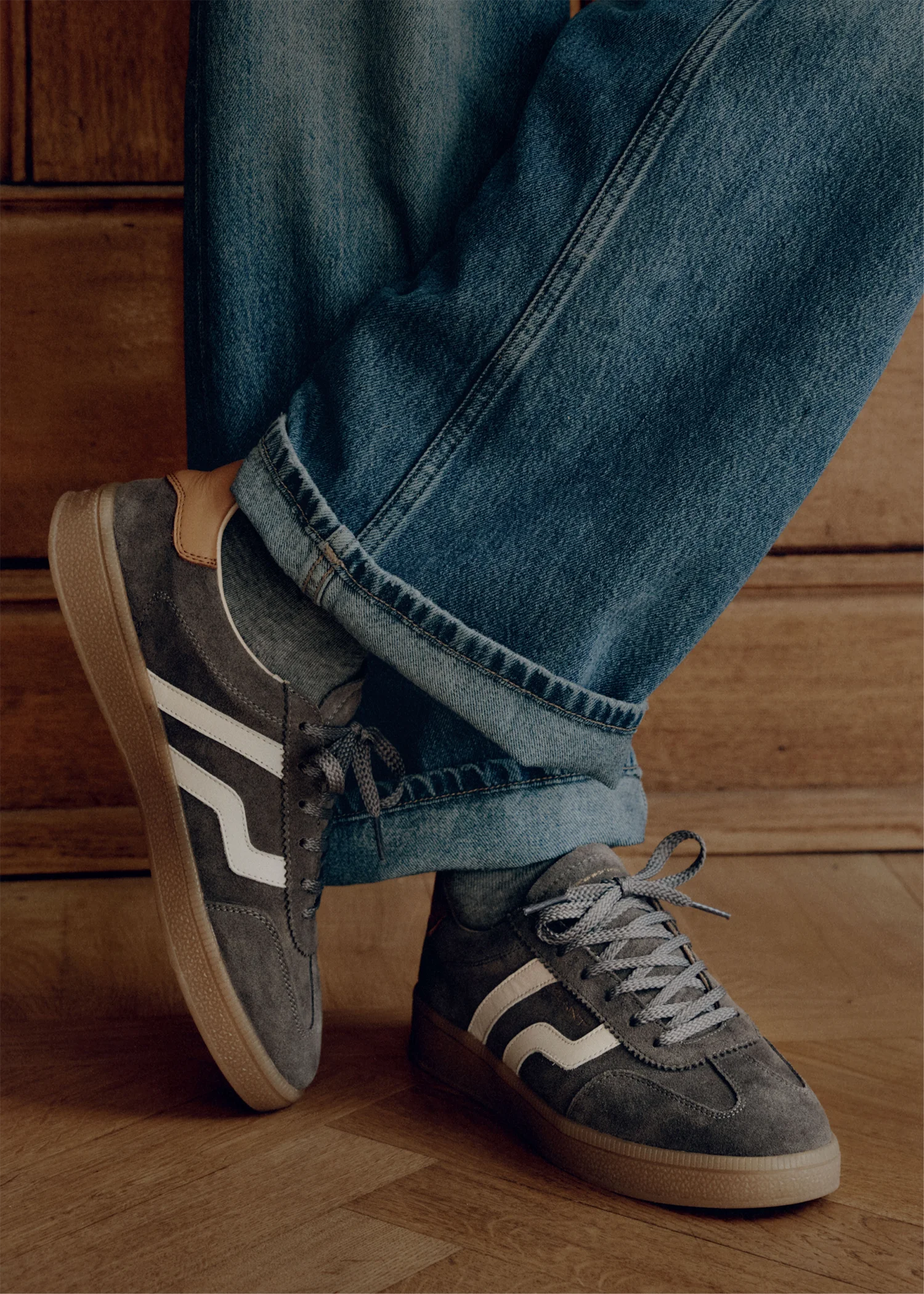 Graue Wildledersneaker mit weißen Streifen, grauen Schnürsenkeln und hellbrauner Sohle, getragen mit blauen Jeans und grauen Socken.