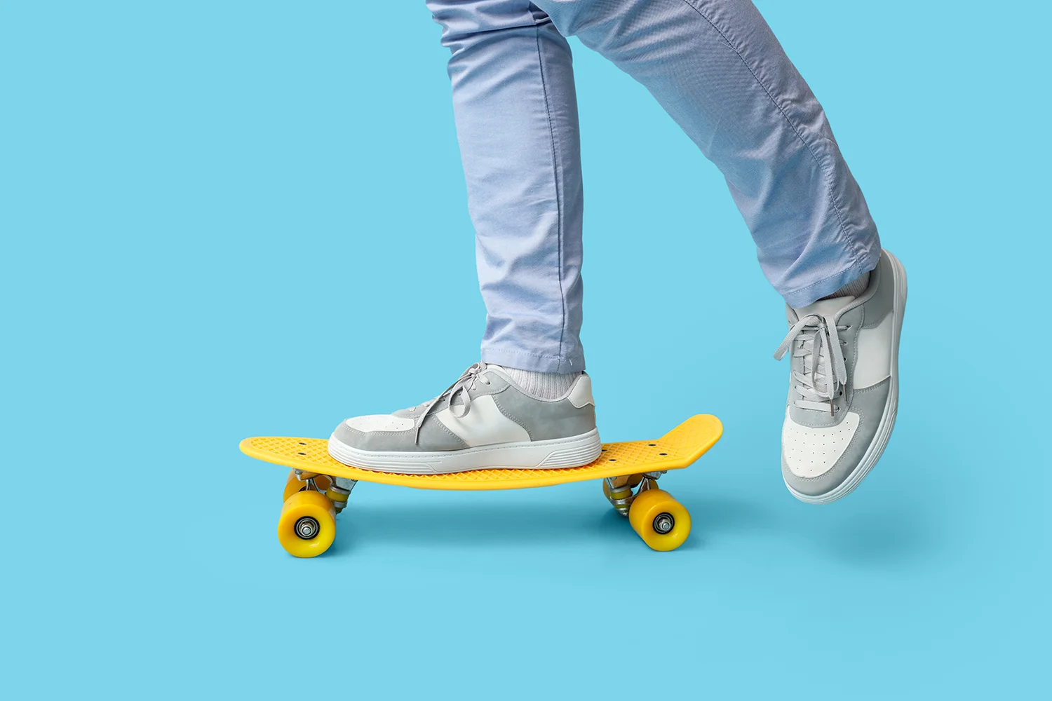 Mann trägt graue Sneaker und hellblaue Jeans, steht auf gelbem Skateboard vor blauem Hintergrund