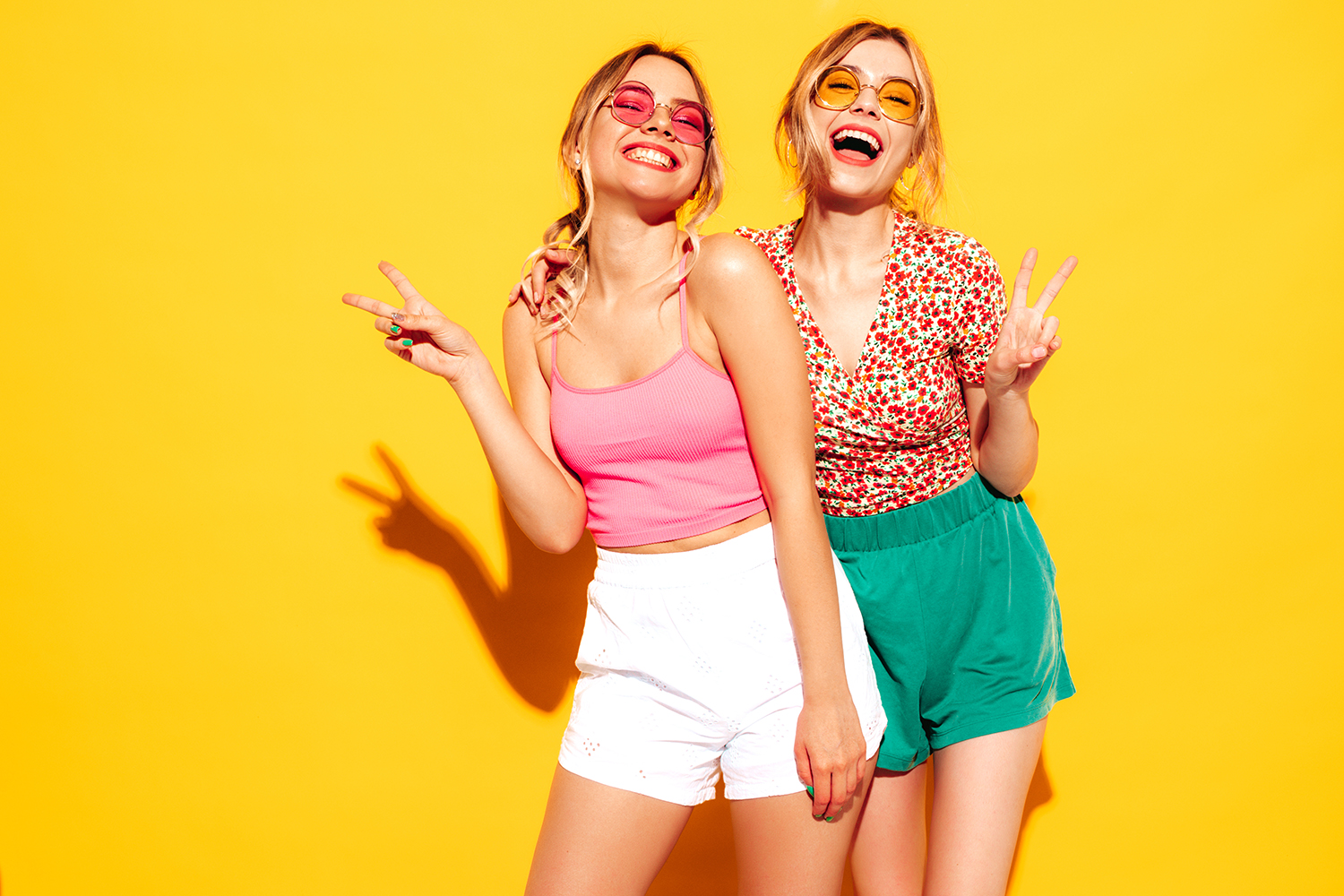 Zwei junge Frauen vor gelbem Hintergrund, eine trägt pinkes Top und weiße Shorts, die andere geblümte Bluse und grüne Shorts, beide zeigen Friedenszeichen
