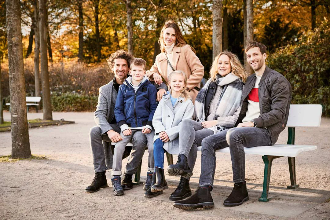 Gruppe von sechs Personen in herbstlicher Parkumgebung, einige sitzen auf einer Bank, andere stehen dahinter, alle tragen Jacken und Stiefel.