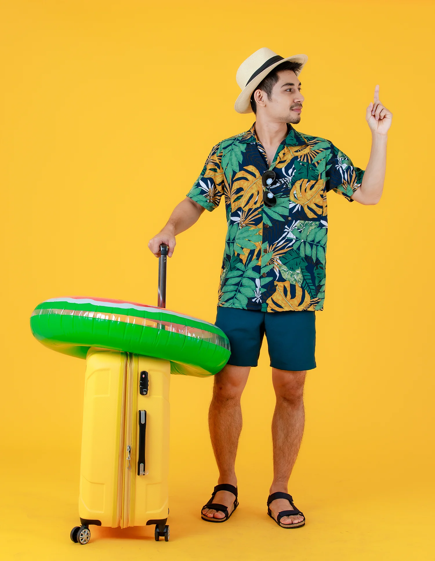 Mann in Hawaii-Hemd, Shorts und Strohhut mit gelbem Koffer und grünem Schwimmring vor gelbem Hintergrund, zeigt nach oben