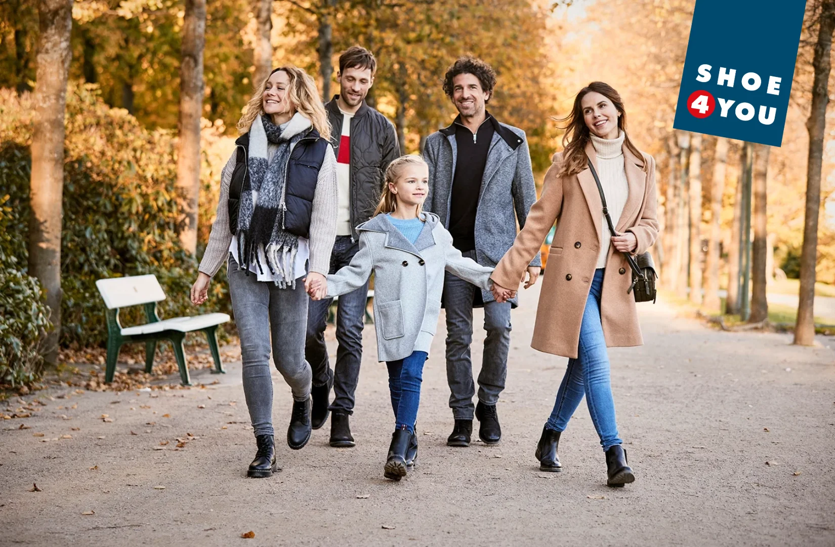 Fünf Personen gehen auf einem von Bäumen gesäumten Weg, zwei halten ein Kind an der Hand, alle tragen Herbstkleidung und Schuhe.