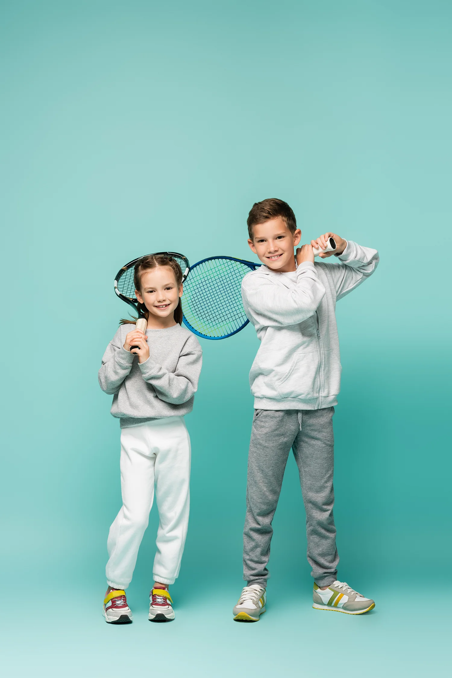 Zwei Kinder in Sportkleidung mit Tennisschlägern, Junge hält Schläger zum Schlag, Mädchen hält Schläger vor sich