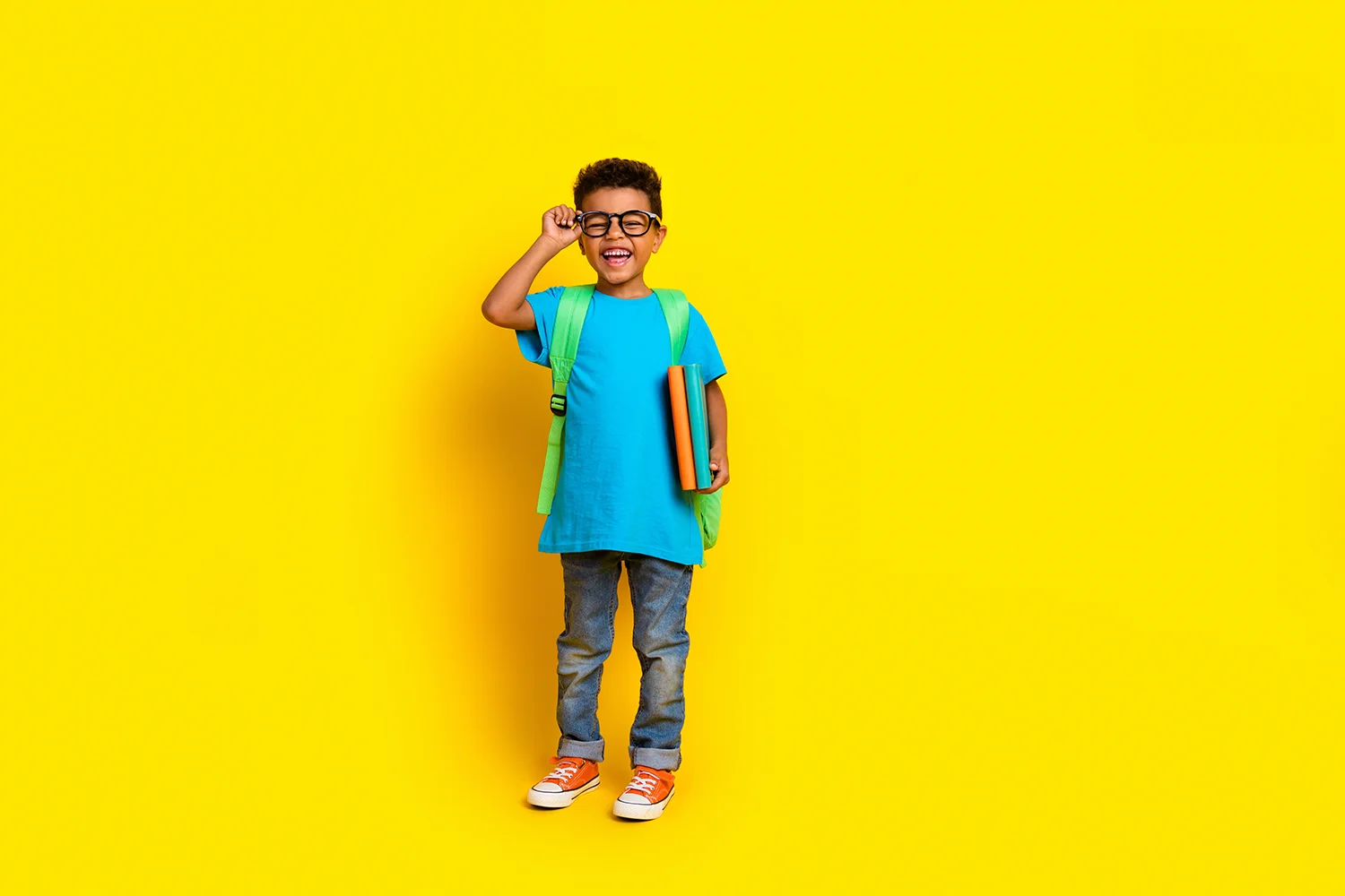 Junge mit Brille, blauem T-Shirt, Jeans und grünem Rucksack hält Bücher vor gelbem Hintergrund
