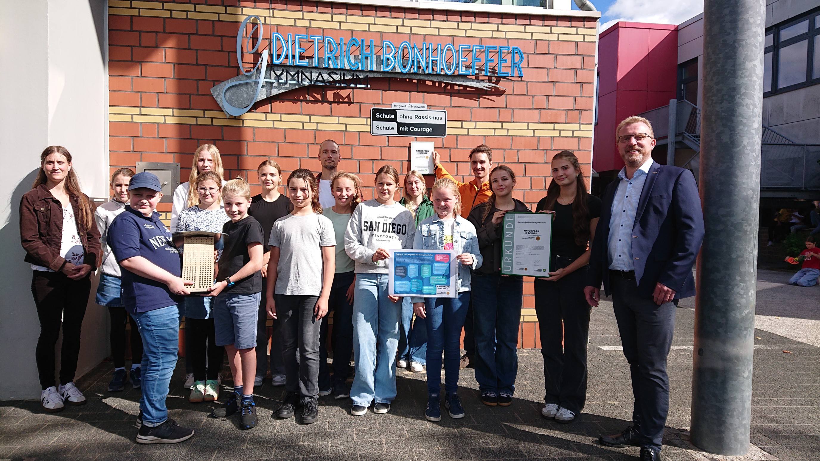 Auf dem Foto sind (von links) Frau Tegeler stellvertretend für den Schulelternrat, verschiedene Aktive der Umwelt-AG der letzten Schuljahre, der für die Koordination am DBG zuständige Lehrer und Leiter der Umwelt-AG, Enno Gesierich (hinten Mitte), Frau Jürgens und Herr Marhoff von der Geschäftsstelle des Zweckverbandes Naturpark Wildeshauser Geest (hinten rechts), Vertreterinnen Schülervertretung (SV) und Schulleiter Florian Schön (rechts) zu sehen.